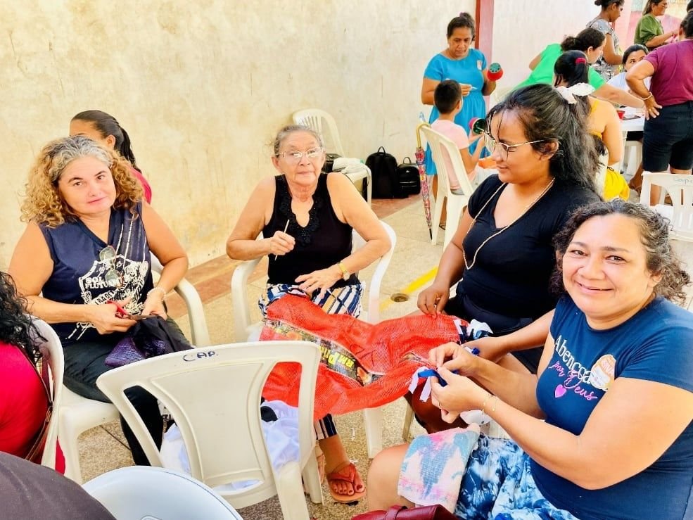 Mulheres sentadas em roda trabalhando juntas em artesanato com sacos de rede vermelha, uma delas sorrindo para a câmera