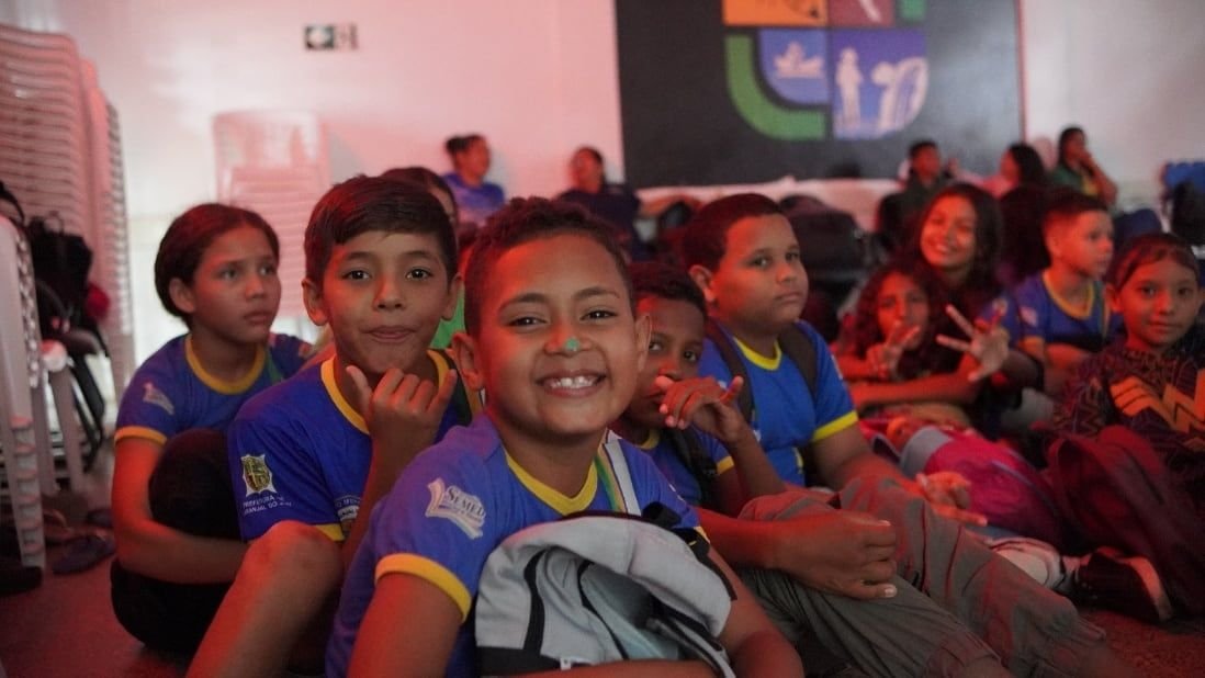 Crianças de uniforme escolar sentadas no chão de uma sala de exibição, sorrindo e fazendo sinais para a câmera