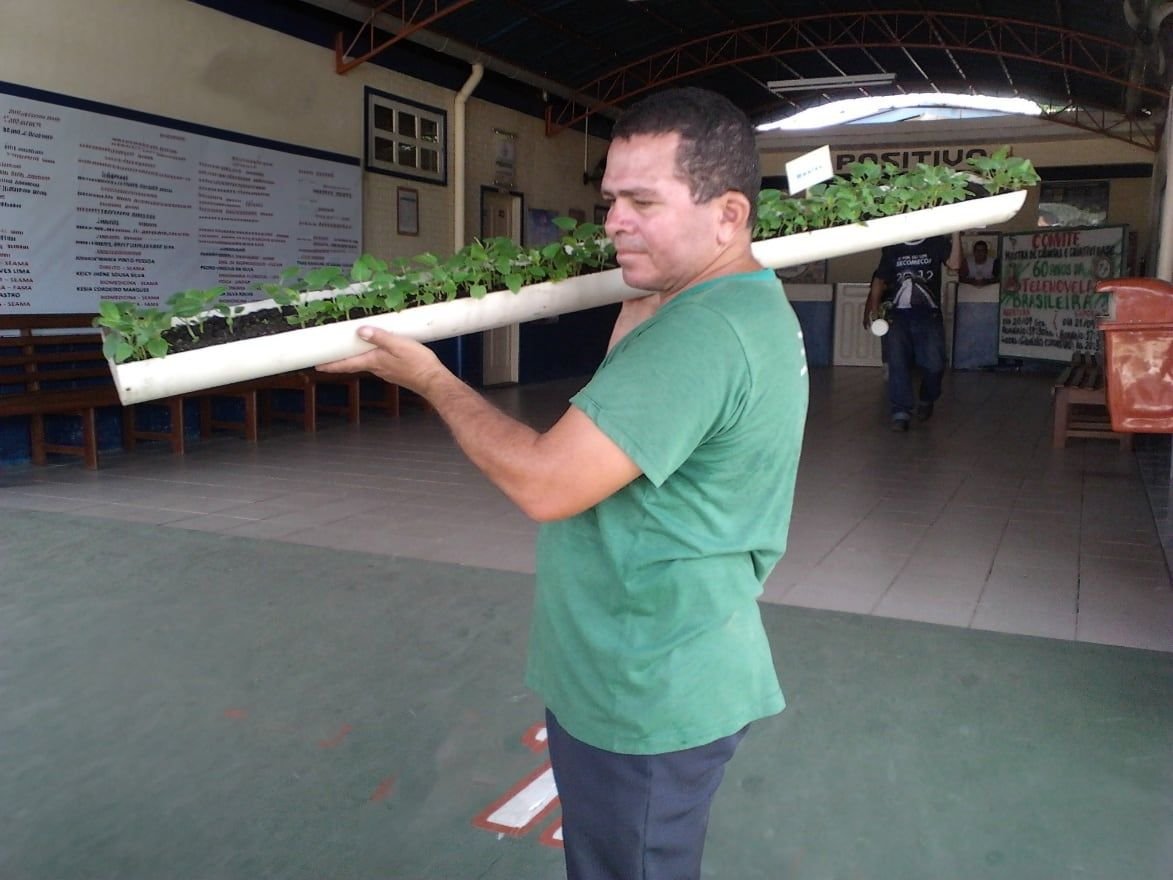 Homem de camiseta verde segurando no ombro um tubo de PVC com mudas de hortaliças