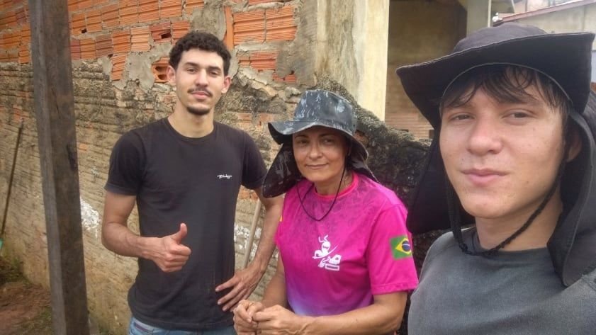 Três pessoas sorrindo para uma selfie ao lado de uma parede de tijolos